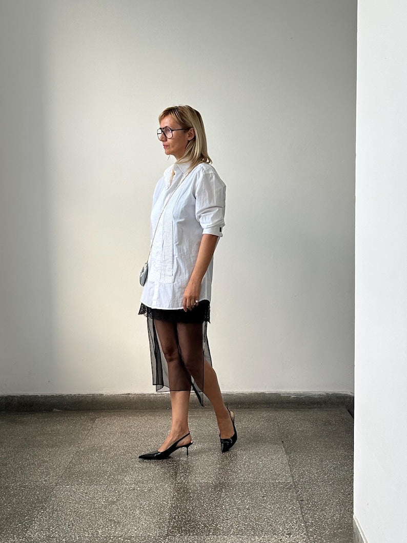 Woman wearing a white shirt and black sheer skirt standing against a plain wall.