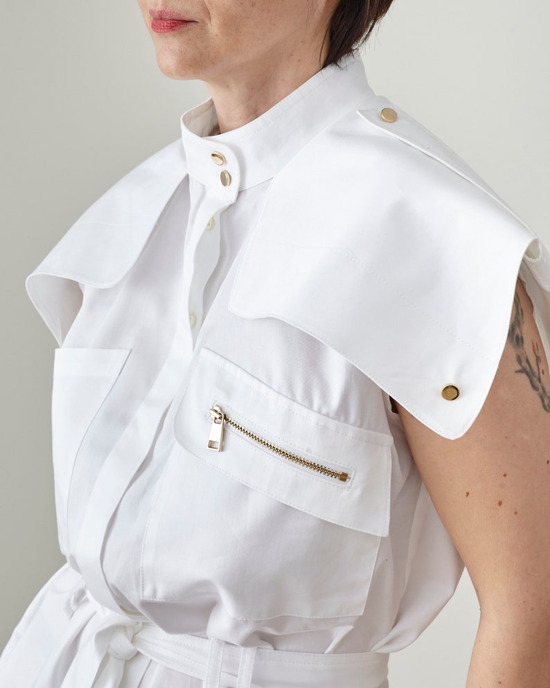 White sleeveless top with gold buttons and zipper chest pocket on a plain background