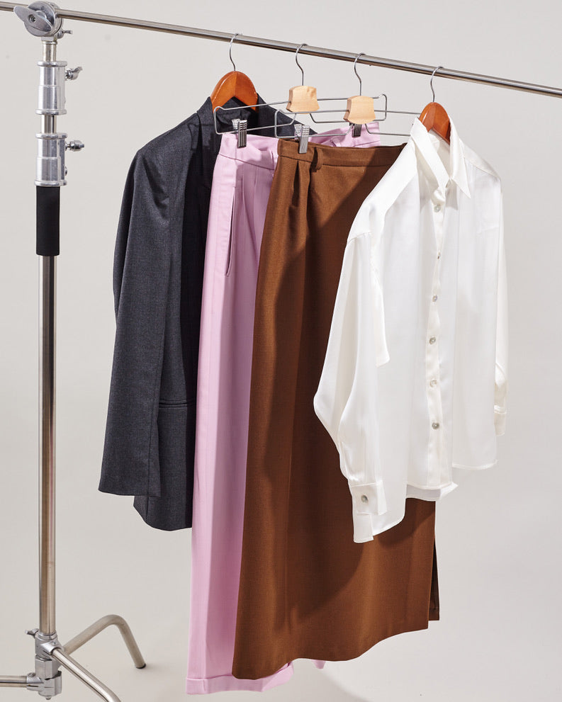 A curated selection of Fine Thee handmade garments on a rolling garment rack, featuring a grey blazer, pink tailored trousers, a brown skirt, and a white silk blouse, representing a sustainable capsule wardrobe.