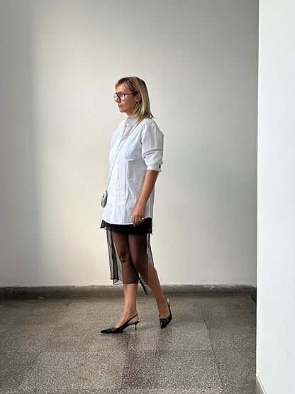 Woman wearing a white shirt and black sheer skirt standing against a plain wall.