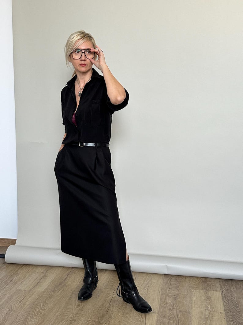 Person wearing a black outfit with a belt and boots, standing against a plain wall.