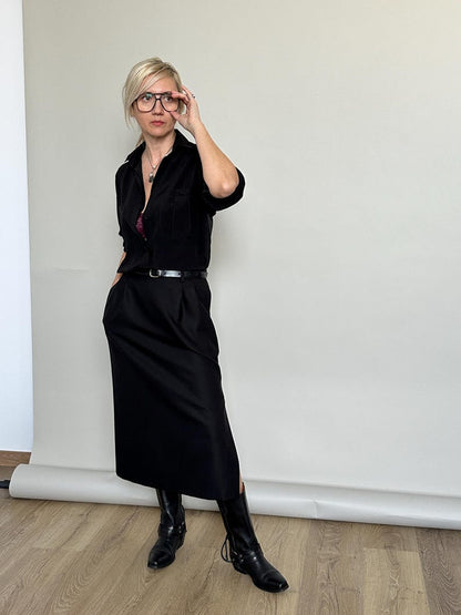 Person wearing a black outfit with a belt and boots, standing against a plain wall.