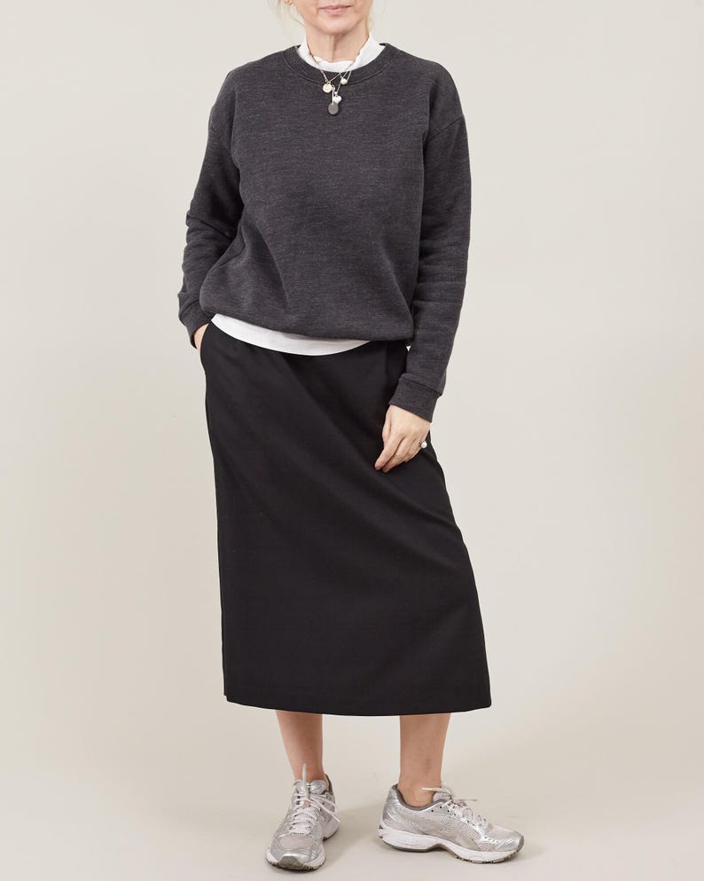 Person wearing a dark gray sweater, white shirt, black skirt, and silver sneakers on a plain background