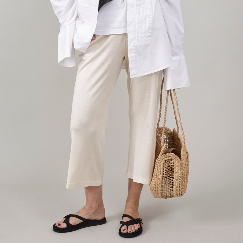 Close-up of Fine Thee ivory white cropped silk jersey pants styled casually with an oversized white shirt, black sandals, and a woven straw tote bag