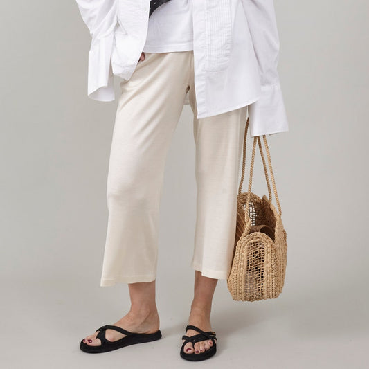 Close-up of Fine Thee ivory white cropped silk jersey pants styled casually with an oversized white shirt, black sandals, and a woven straw tote bag