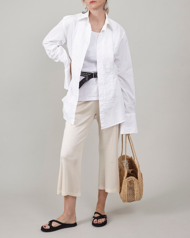 Full-body studio photograph of Fine Thee ivory cropped silk jersey pants on a woman with clean, unmarked skin. She is styled with an oversized pleated white shirt, black belt, black sandals, large grey earrings, and holding a woven straw tote bag against a neutral grey background