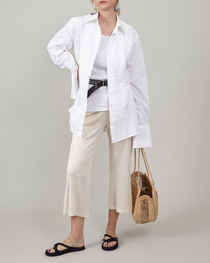 Full-body studio photograph of Fine Thee ivory cropped silk jersey pants on a woman with clean, unmarked skin. She is styled with an oversized pleated white shirt, black belt, black sandals, large grey earrings, and holding a woven straw tote bag against a neutral grey background