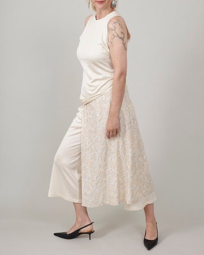 Side-angle shot of Fine Thee ivory cropped silk jersey pants layered under a cream asymmetric dress, paired with black kitten heel pumps.