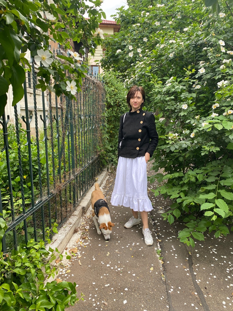 Woman in black jacket and white skirt standing with a dog on a path surrounded by greenery