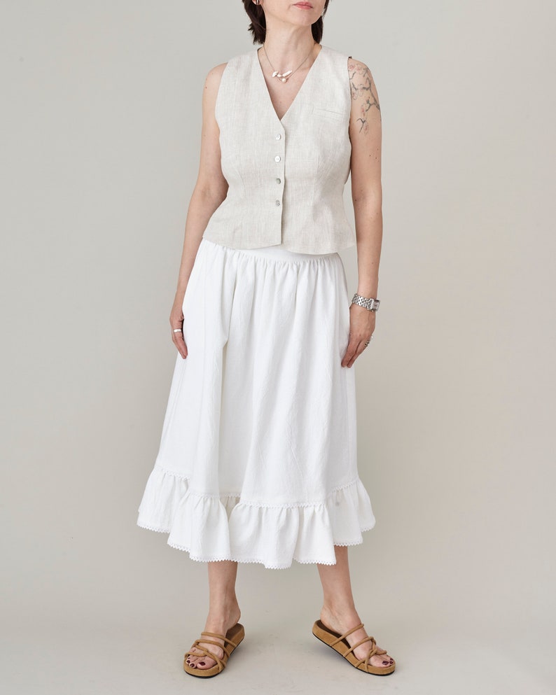 Woman wearing a cream tailored vest and a white gathered skirt with a plain background
