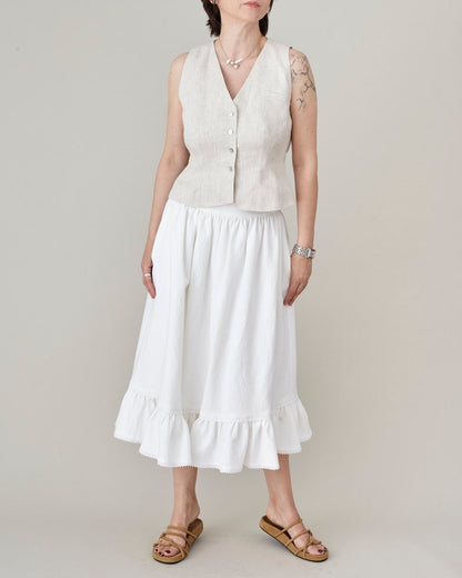 Woman wearing a cream tailored vest and a white gathered skirt with a plain background