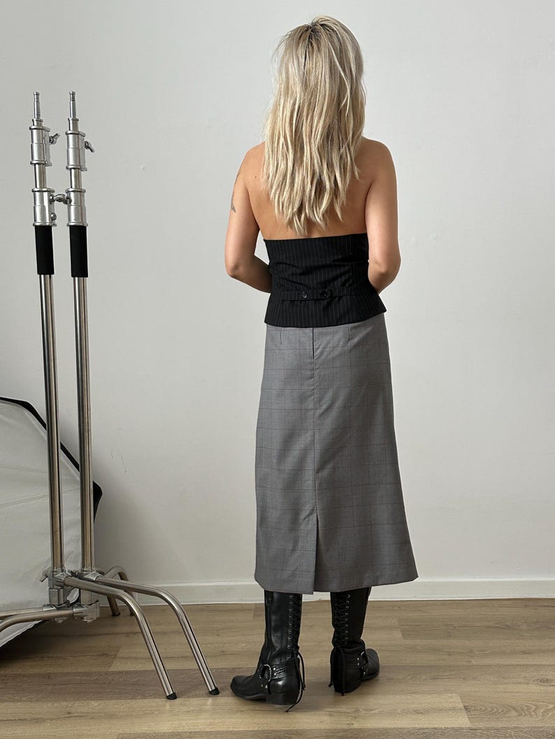 Woman wearing a black top and gray skirt standing in a room with a white wall and wooden floor.