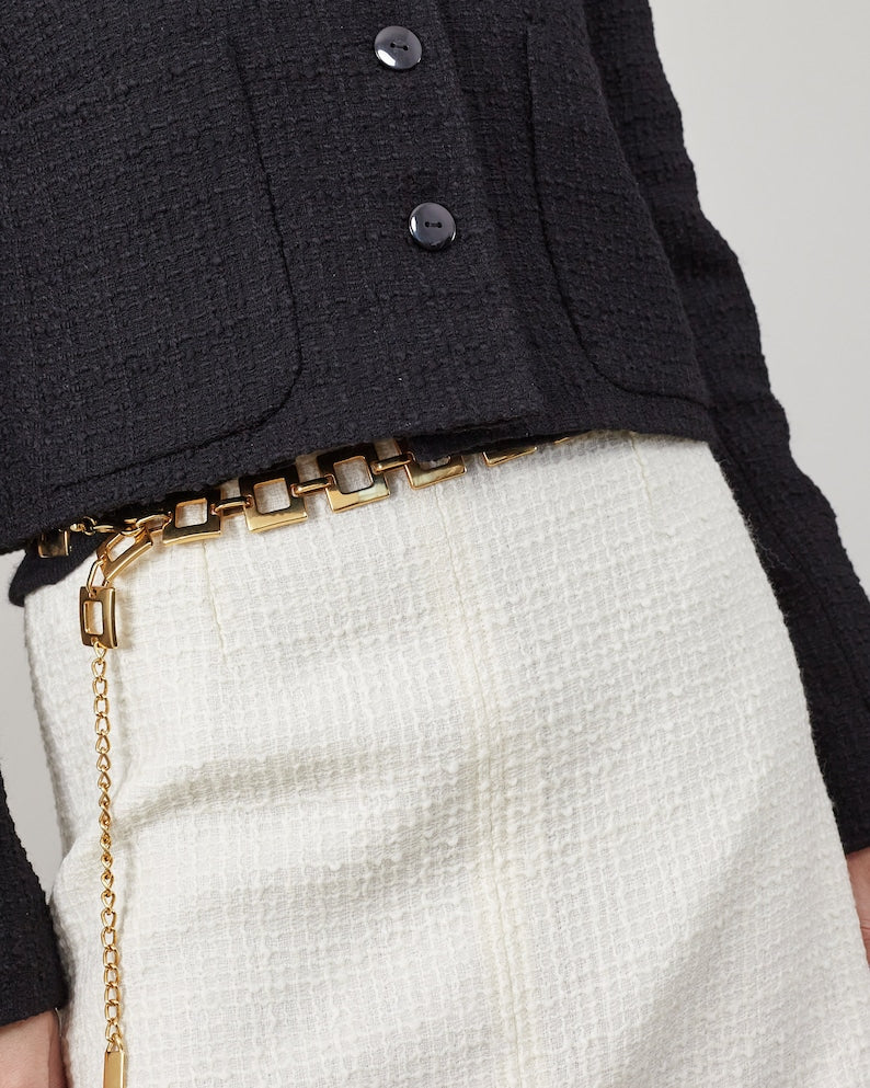 Close-up of a wool boucle black jacket with gold metallic belt and white mini skirt on a neutral background