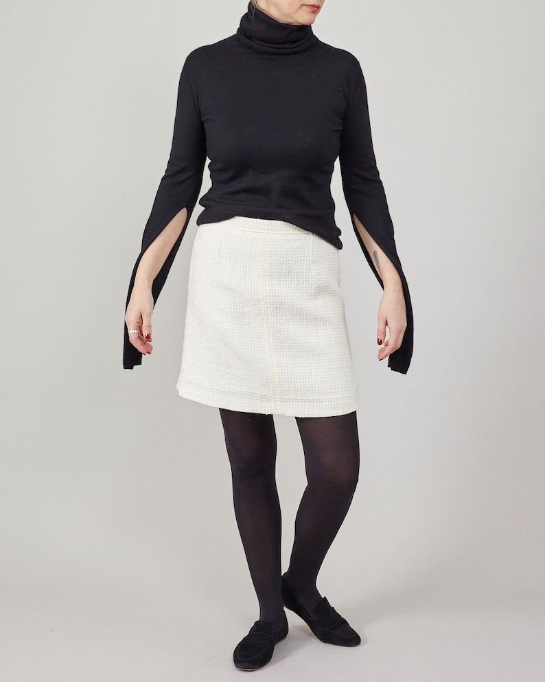 Person wearing a black turtleneck, white skirt, and black loafers on a plain background