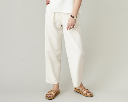 Close-up of handmade ivory raw silk tapered trousers with pleated high waist and cropped hem, styled with tan strappy sandals, by Fine Thee