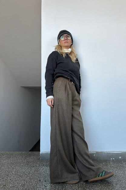 Person wearing a black sweater and brown wide-leg pants standing against a white wall.