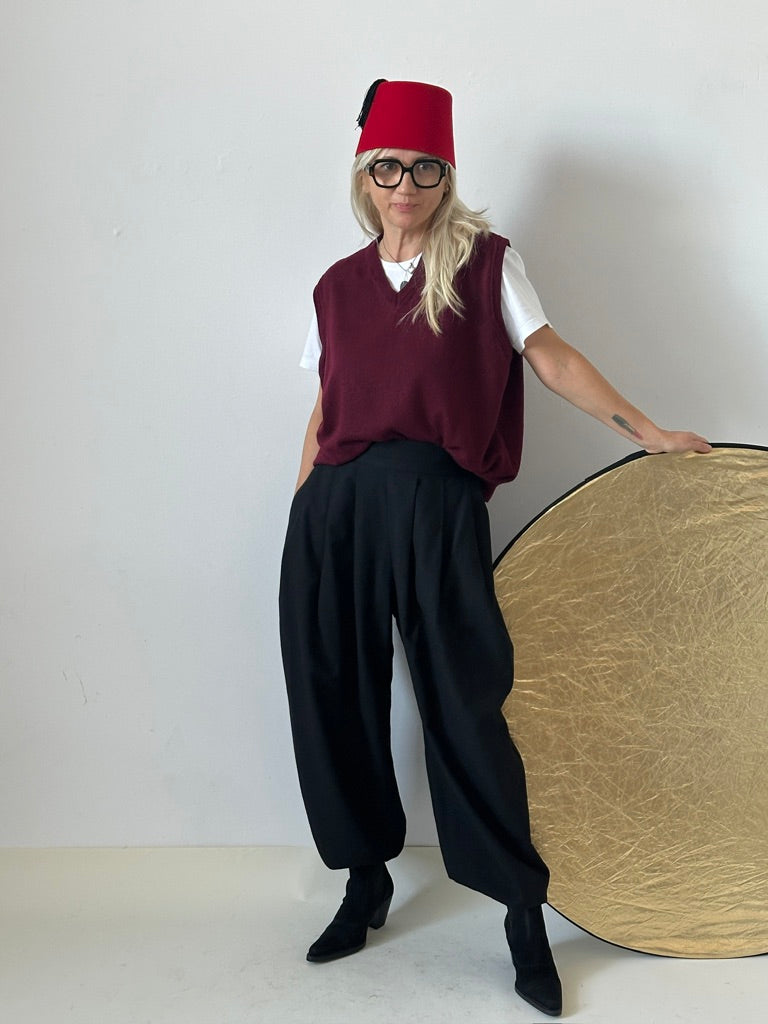 A stylish woman in the atelier posing in voluminous high-waisted black wool tailored trousers with pleats, a burgundy sweater vest over a white t-shirt, wearing a red fez hat and glasses, with a gold reflector disk on the side, showcasing Fine Thee's unique artisanal tailoring.