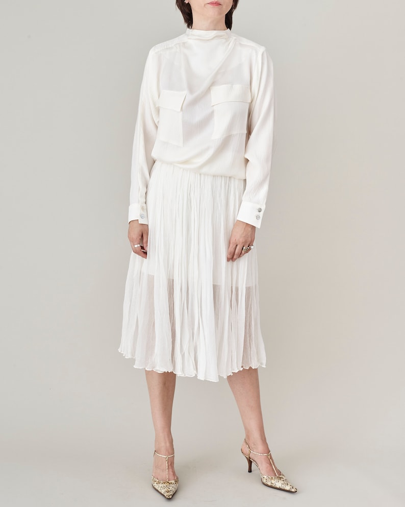 Person wearing a white long-sleeve top and pleated skirt on a plain background