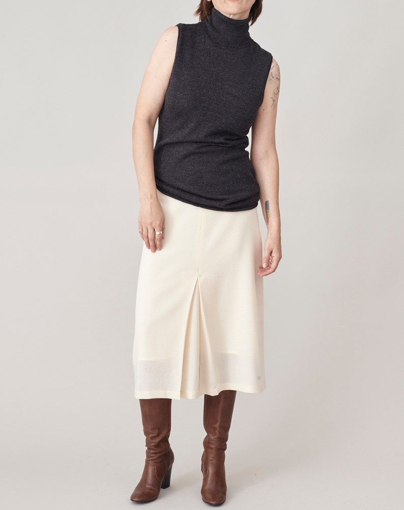 Person wearing a dark sleeveless top, cream skirt, and brown boots on a plain background
