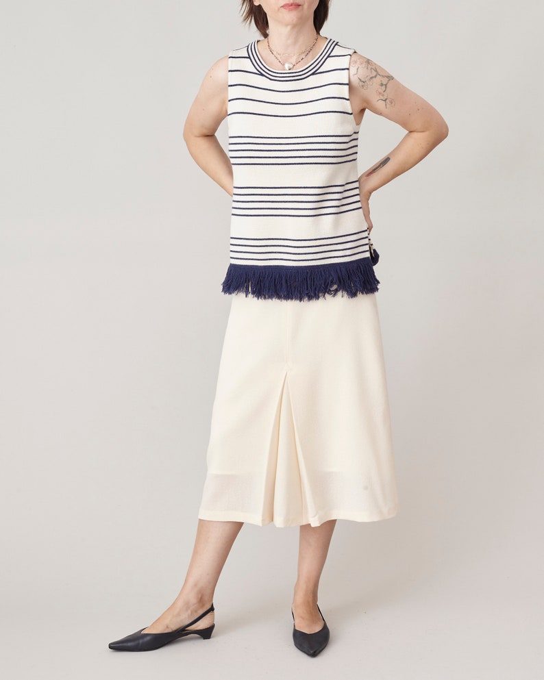 Woman wearing a sleeveless striped top with a fringed hem and a cream skirt on a plain background