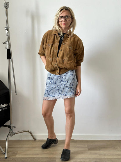 Woman wearing a brown jacket and floral skirt standing against a plain wall.