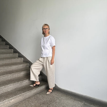 Person wearing a white top and beige pants standing on a staircase against a plain wall.