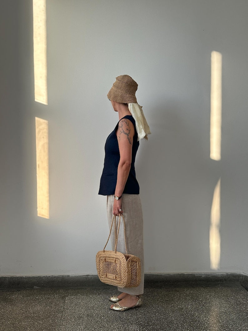 Person wearing a navy top and barrel leg beige pants, holding a woven bag against a plain wall with light streaming in