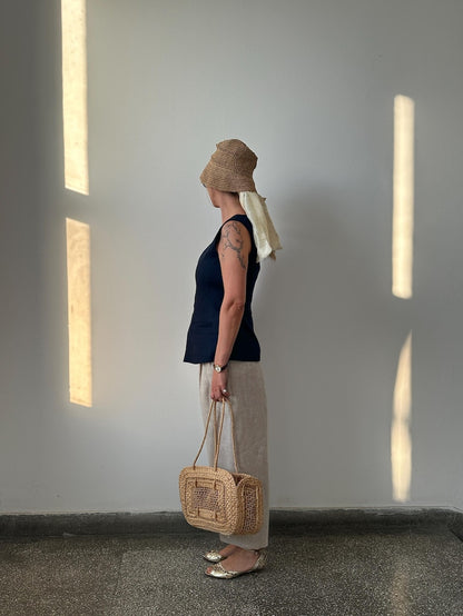 Person wearing a navy top and barrel leg beige pants, holding a woven bag against a plain wall with light streaming in