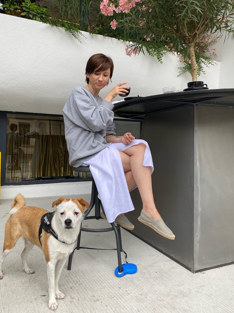 Woman wearing a white skirt, sitting at a bar with a dog next to her, surrounded by plants and a white wall.
