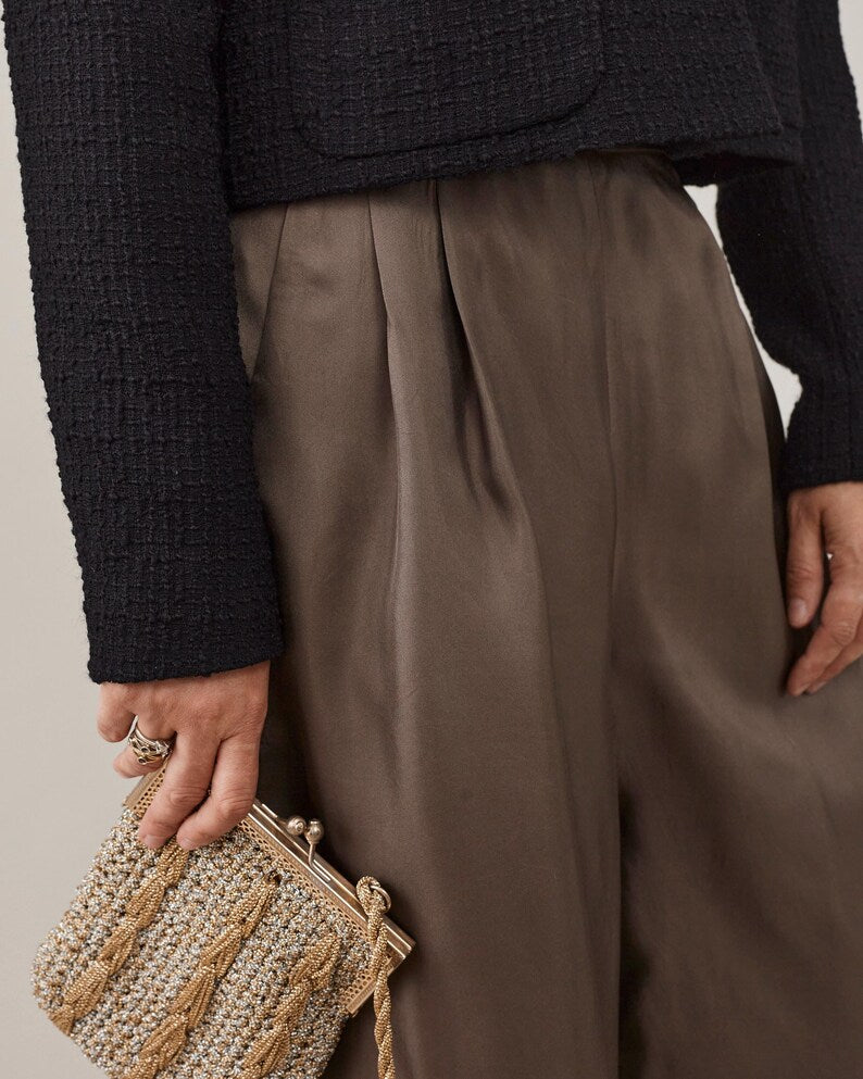 Person wearing a black textured crop jacket and taupe silk wide-leg pants, holding a beaded clutch.