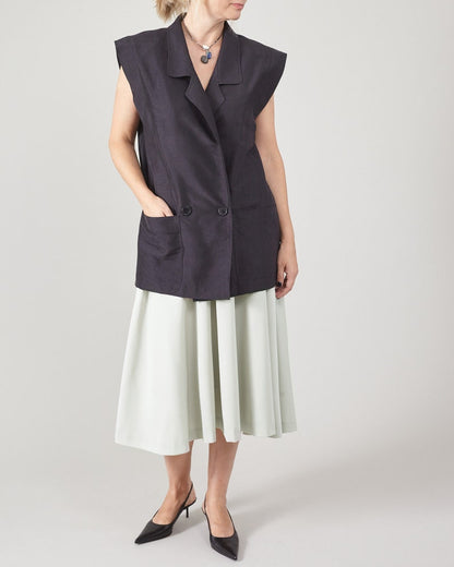 Woman wearing a dark sleeveless jacket over a light mint skirt on a plain background