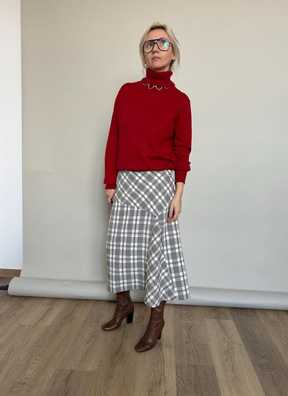 Woman wearing a red sweater and plaid skirt standing against a plain wall.