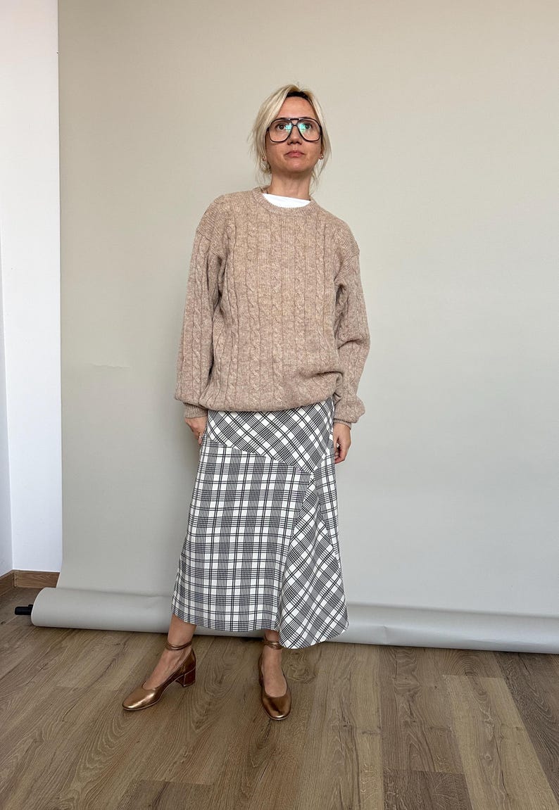 Woman wearing a beige sweater and plaid skirt standing against a plain wall.