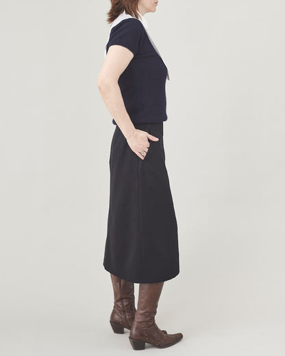 Person wearing a navy sweater with a white collar, black midi skirt and brown boots on a plain background