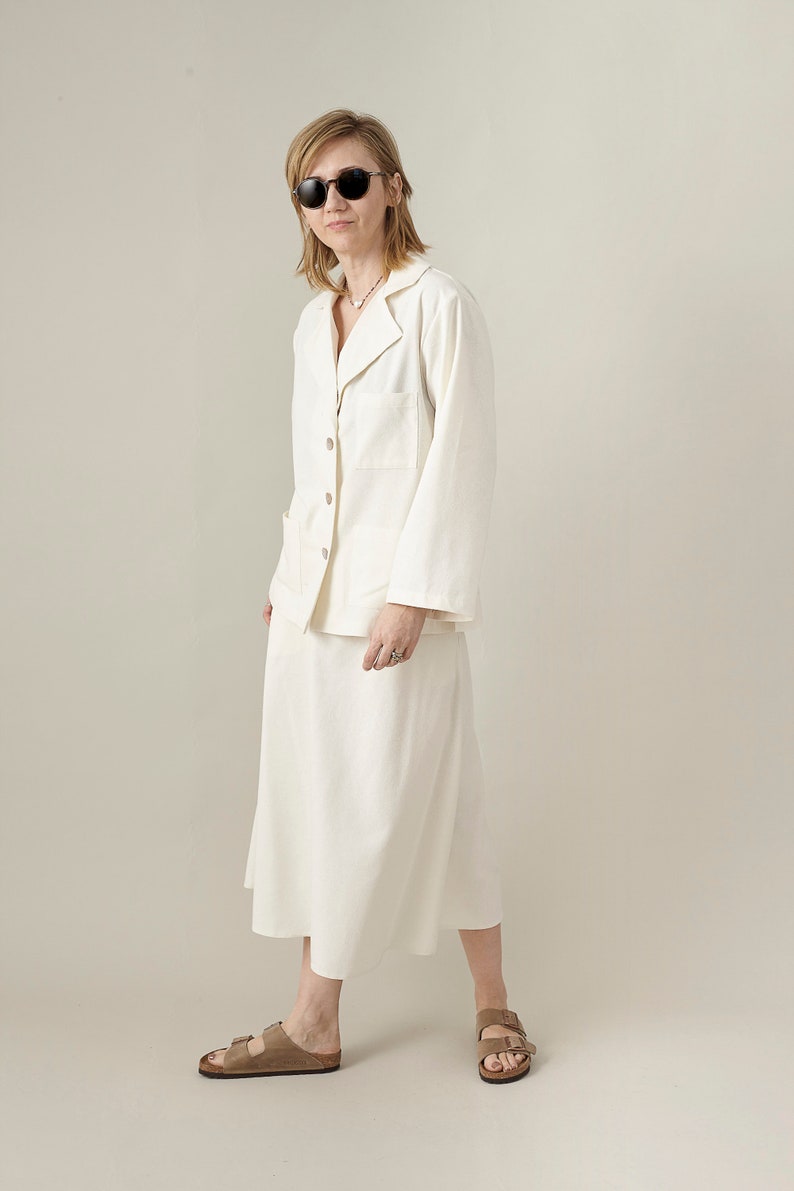 Woman wearing a white outfit, blouse and matching midi skirt with sunglasses on a plain background