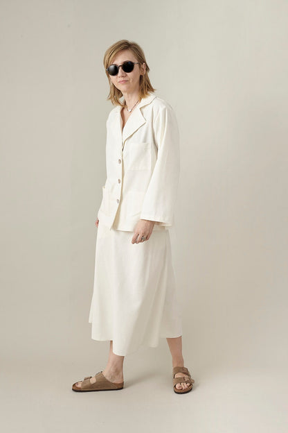 Woman wearing a white outfit, blouse and matching midi skirt with sunglasses on a plain background