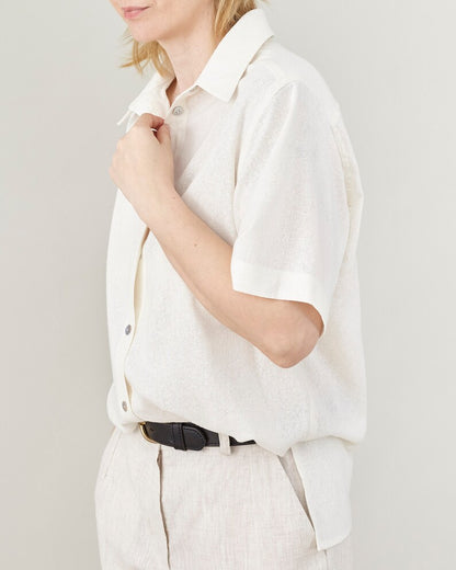Person wearing a white short sleeve shirt with mother of pearl buttons, oversized fit and side slit, tucked into linen pants.