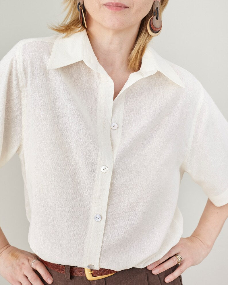 Person wearing an off white button-up shirt in a lightweight, slightly sheer fabric a plain background