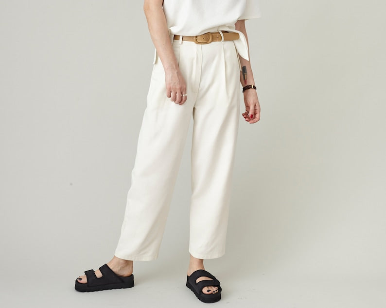 Close-up view of pleated raw silk tapered trousers in off-white, featuring a comfortable high-waisted fit, belt loops, and minimalist styling with black slide sandals.