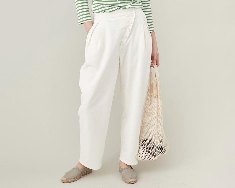 Person wearing white high waisted pants with pleats and a green striped shirt, holding a net bag against a plain background