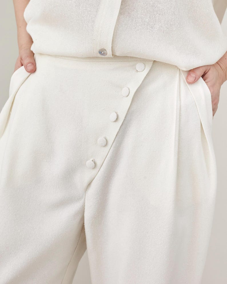 Close-up of a person wearing white high waisted pleated pants with fabric covered buttons flap 