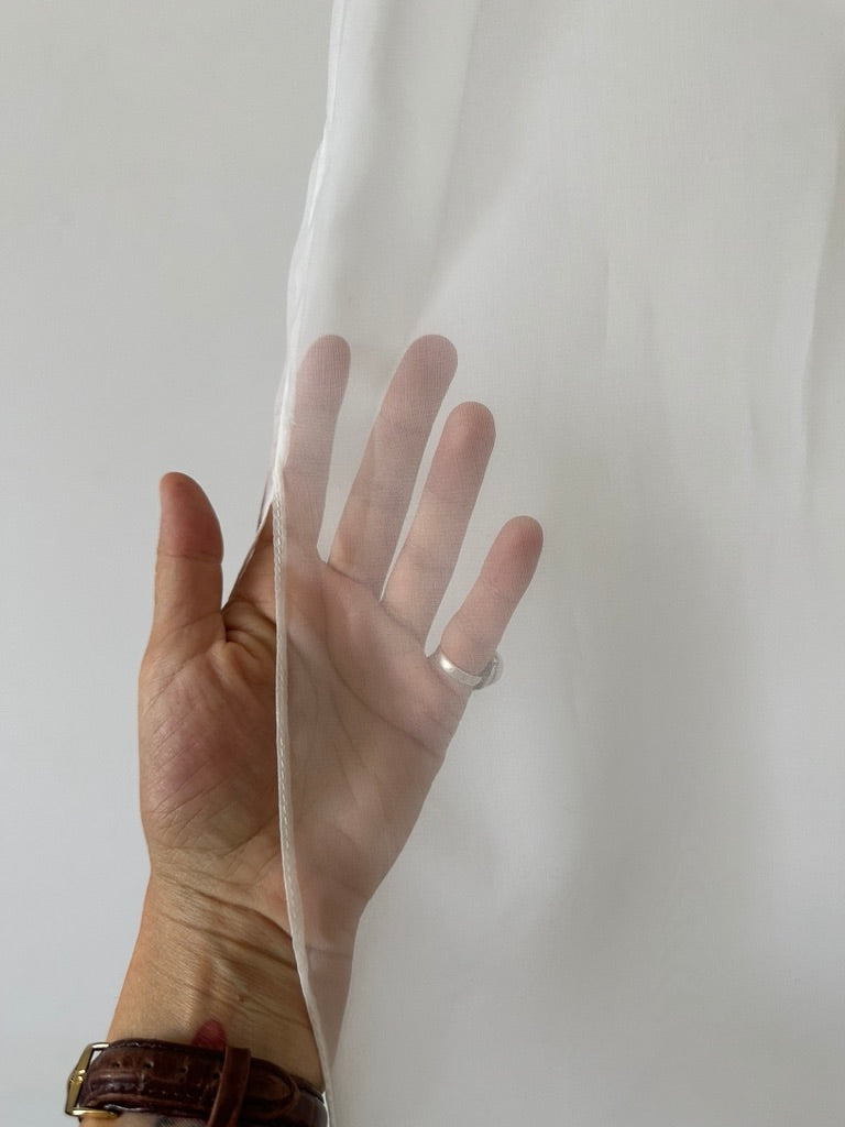 A close-up photograph showing a woman's hand with a ring holding the hem of a sheer, ethereal white silk organza fabric, highlighting the texture and quality of the premium natural material used by Fine Thee.