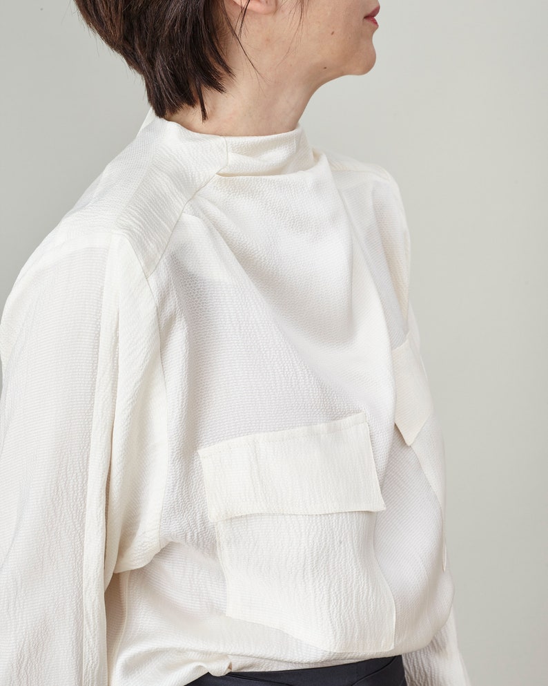 Person wearing a white blouse with a high collar and patch pockets on a plain background