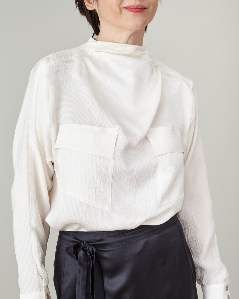 Person wearing a white blouse with a high collar and black skirt against a gray background