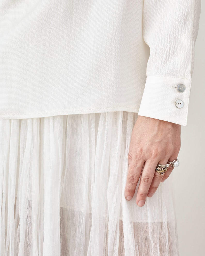 Close-up of a person wearing a white shirt and sheer white skirt with a pearl ring on a plain background