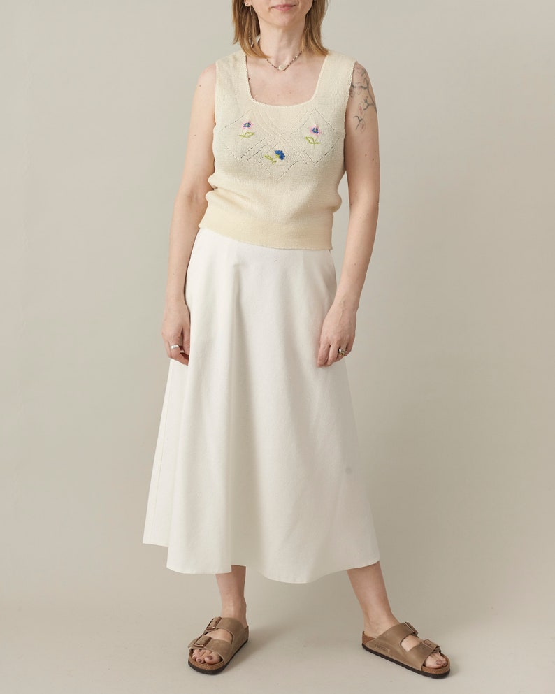 Person wearing a light-colored sleeveless top with floral patterns and a white skirt on a plain background