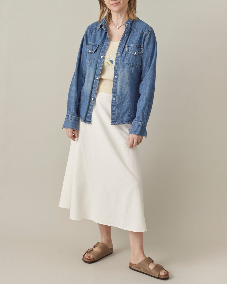 Person wearing a denim jacket, white skirt, and sandals on a plain background