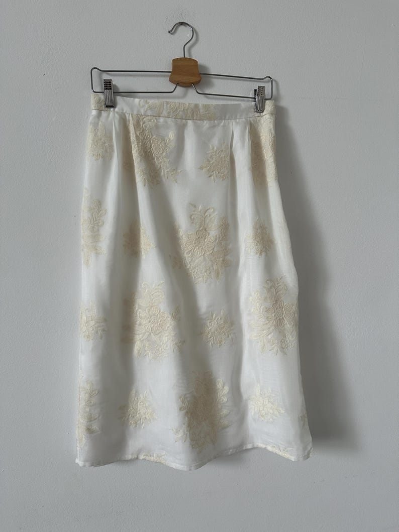 White skirt with ivory floral embroidery on a hanger against a gray wall