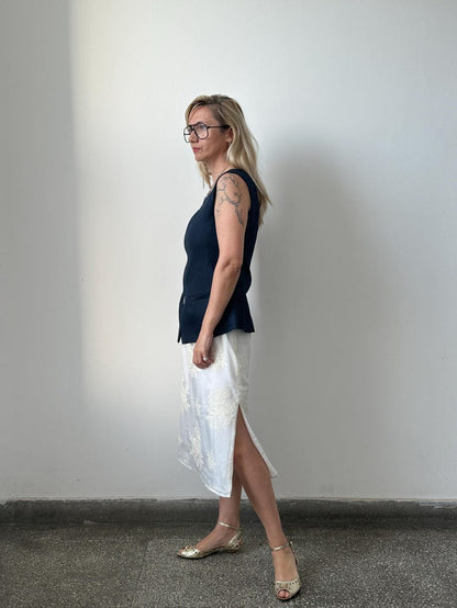 Woman wearing a navy top and white skirt against a plain wall.