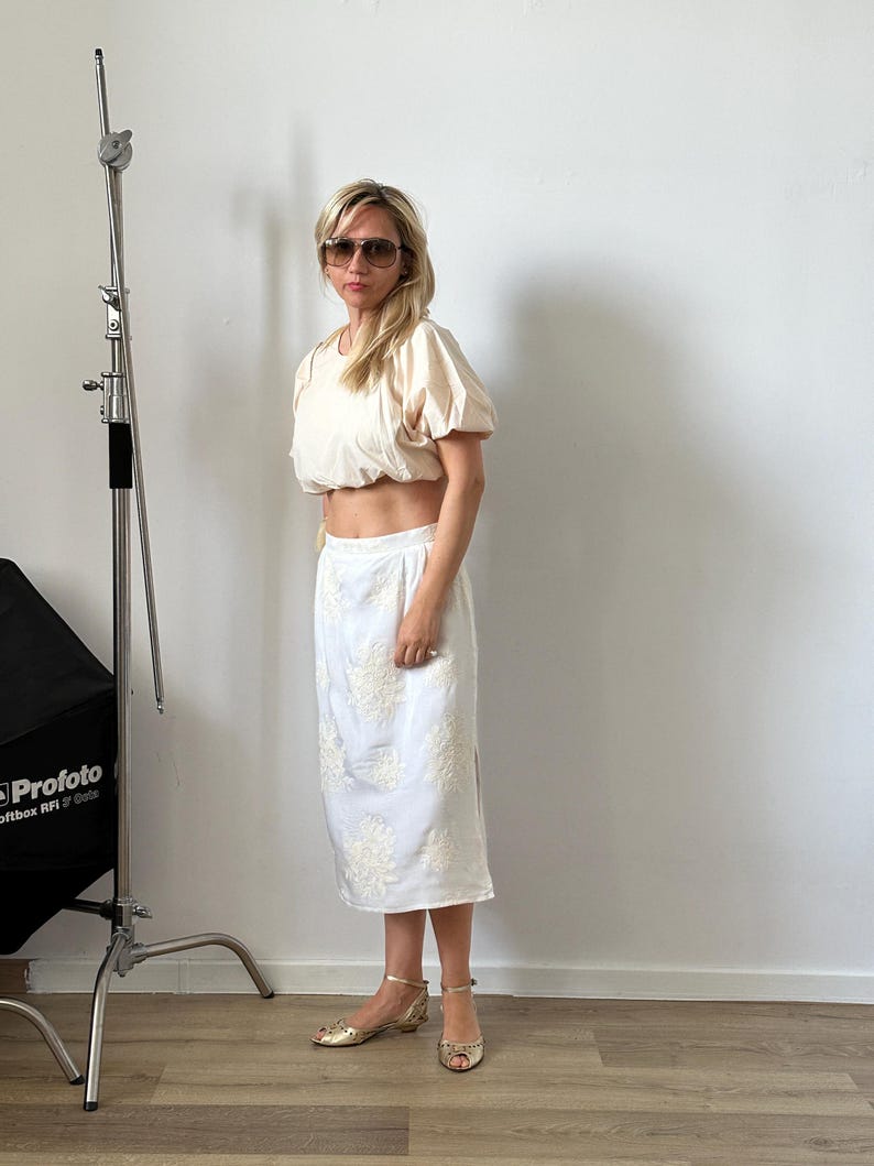 Woman in a white outfit standing in front of a white wall with photography equipment visible.
